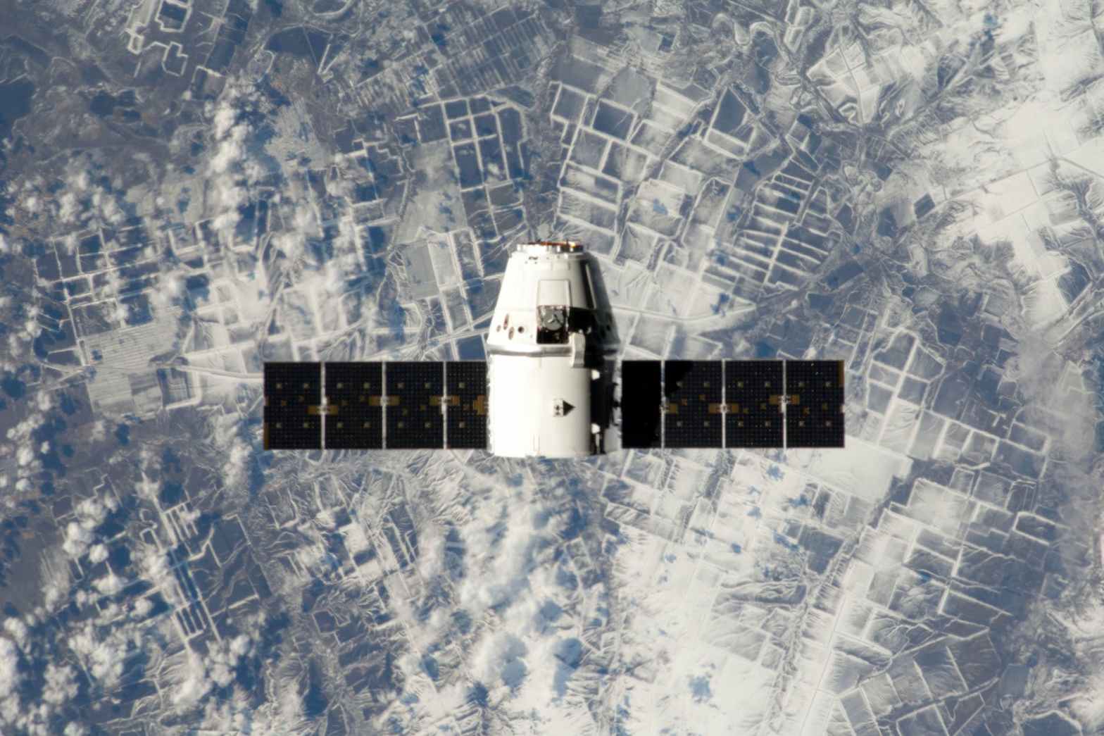 A SpaceX dragon capsule in Earth Orbit. The solar panels extend from the left and right sides of the craft as the Earth is visible below.